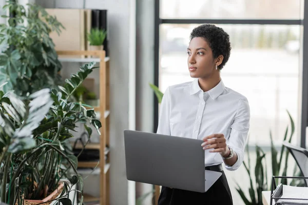 Pensativa mujer de negocios afroamericana con portátil mirando hacia otro lado, mientras que de pie cerca de plantas verdes en la oficina moderna - foto de stock
