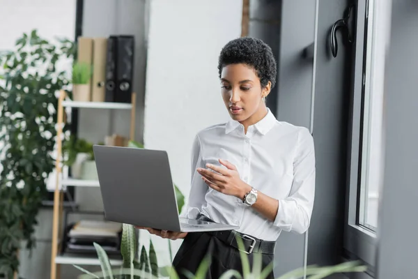 Joven mujer de negocios afroamericana en blusa blanca señalando con la mano durante el chat de vídeo en el ordenador portátil en la oficina - foto de stock