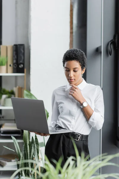 Молодая африканская американская бизнесвумен думает и смотрит на ноутбук, стоя в офисе — стоковое фото