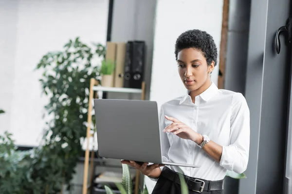 Elegante mujer de negocios afroamericana en blusa blanca señalando con el dedo durante la videollamada en el ordenador portátil en la oficina - foto de stock