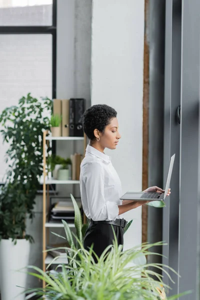 Vista lateral de la mujer de negocios afroamericana en blusa blanca de pie con computadora portátil en la oficina contemporánea - foto de stock