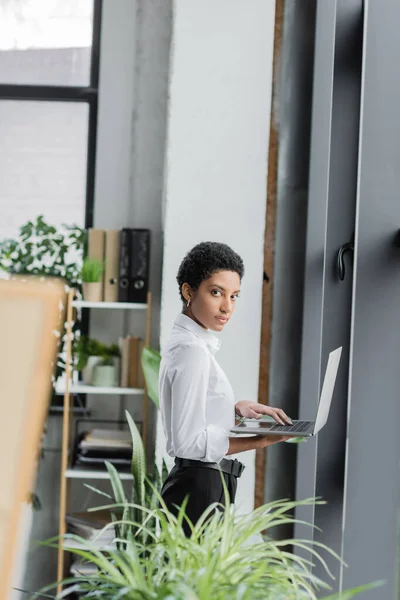 Stylish african american businesswoman in white blouse looking at camera while standing with laptop in modern office — Stock Photo