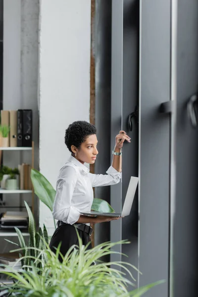 Вдумчивая американская бизнесвумен с ноутбуком, смотрящая в сторону окон в современном офисе — стоковое фото