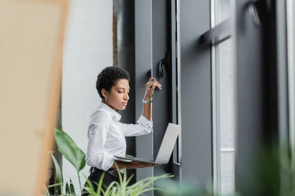 Élégante femme d'affaires afro-américaine debout près des fenêtres dans le bureau et regardant ordinateur portable — Photo de stock
