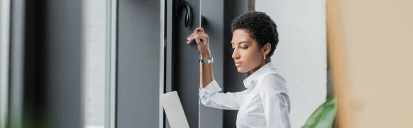 Vista lateral de la joven mujer de negocios afroamericana en blusa blanca que sostiene el ordenador portátil cerca de ventanas en la oficina, pancarta - foto de stock