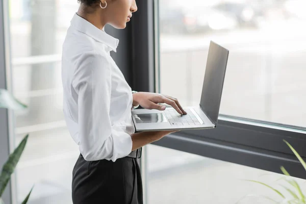 Vista recortada de la mujer de negocios afroamericana en blusa blanca usando computadora portátil con pantalla en blanco cerca de la ventana en la oficina - foto de stock