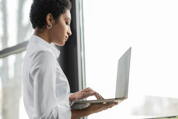 Vista lateral de la mujer de negocios afroamericana en blusa blanca de pie cerca de la ventana en la oficina y utilizando el ordenador portátil - foto de stock