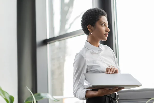 Pensativa mulher de negócios afro-americana segurando laptop e olhando para longe perto da janela no escritório — Fotografia de Stock