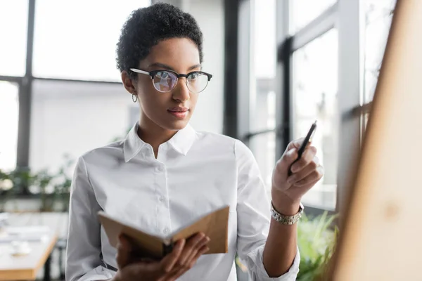 Jovem empresária afro-americana em óculos segurando bloco de notas e apontando com caneta em primeiro plano desfocado no escritório — Fotografia de Stock