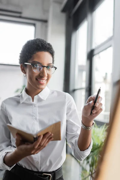 Mujer de negocios afroamericana positiva en gafas con estilo sosteniendo cuaderno y señalando con pluma en la oficina - foto de stock