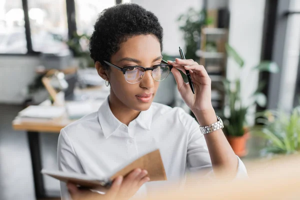 Mujer de negocios afroamericana en blusa blanca ajustando gafas mientras sostiene bloc de notas y bolígrafo en la oficina - foto de stock