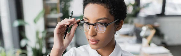 Jeune femme d'affaires afro-américaine tenant un stylo et ajustant des lunettes élégantes au bureau, bannière — Photo de stock