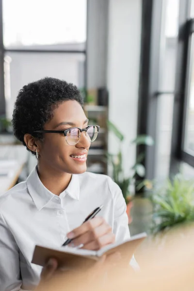 Жизнерадостная африканская американская бизнесвумен в стильных очках и белой блузке, пишущая в блокноте и глядя в сторону в офисе — стоковое фото