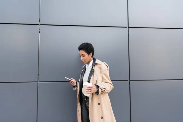 Молодая африканская американская предпринимательница в бежевом плаще, обменивающаяся сообщениями на смартфоне и держащая бумажную чашку возле серой стены на улице — стоковое фото