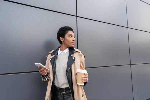 Mulher de negócios afro-americana em casaco de trincheira elegante segurando copo de papel e telefone celular enquanto olha para longe perto da parede cinza na rua — Fotografia de Stock