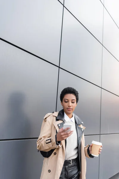 Junge afrikanisch-amerikanische Geschäftsfrau in schickem Trenchcoat mit Pappbecher und Botschaften auf dem Smartphone in der Nähe einer grauen Wand auf der städtischen Straße — Stockfoto