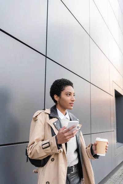 Femme d'affaires afro-américaine en trench coat beige debout avec tasse en papier et smartphone près du mur gris sur la rue de la ville — Photo de stock