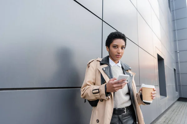 Donna d'affari afro-americana alla moda con tazza usa e getta e smartphone guardando la fotocamera vicino al muro grigio sulla strada in città — Foto stock