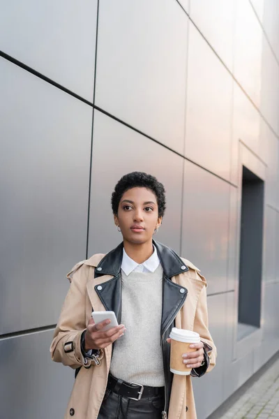 Junge afrikanisch-amerikanische Geschäftsfrau in modischem Trenchcoat hält Smartphone und Kaffee in der Nähe einer grauen Mauer auf der städtischen Straße — Stockfoto