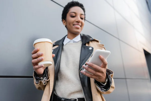 Blurred african american businesswoman with coffee to go smiling and messaging on cellphone near grey wall on urban street — Stock Photo