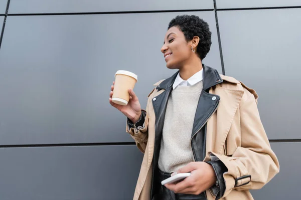 Happy african american businesswoman in stylish trench coat holding takeaway drink and mobile phone near wall on urban street — Stock Photo