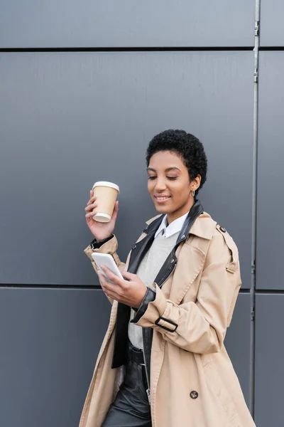 Mujer de negocios afroamericana positiva en gabardina beige charlando en un teléfono inteligente y sosteniendo bebida para llevar cerca de la pared gris en la calle - foto de stock