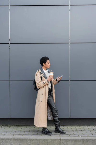 Afrikanisch-amerikanische Geschäftsfrau in Trenchcoat und Lederstiefeln hält Einwegbecher in der Hand und plaudert auf der Straße mit dem Handy — Stockfoto