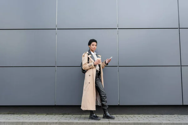 Piena lunghezza di donna afroamericana in trench beige in piedi con drink da asporto e smartphone vicino al muro grigio sulla strada — Foto stock
