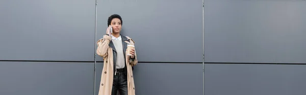Donna d'affari afroamericana alla moda che tiene la tazza usa e getta e parla sul telefono cellulare vicino al muro grigio sulla strada della città, banner — Foto stock
