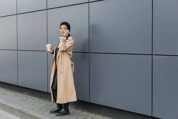 Femme d'affaires afro-américaine en trench-coat beige debout avec boisson à emporter et appelant sur smartphone près du mur gris en ville — Photo de stock