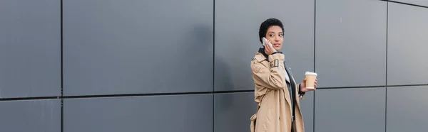 Femme d'affaires afro-américaine en trench coat beige debout avec boisson à emporter et parler sur smartphone dans la rue, bannière — Photo de stock