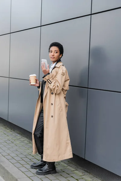In voller Länge trendige afrikanisch-amerikanische Geschäftsfrau mit Coffee to go und Handy, das in der Nähe einer grauen Mauer auf der städtischen Straße wegschaut — Stockfoto