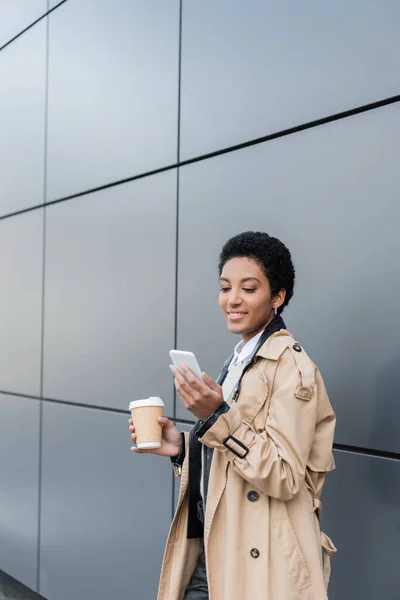 Alegre mujer de negocios afroamericana en gabardina beige sosteniendo café en taza de papel y charlando en el teléfono inteligente cerca de la pared gris en la ciudad - foto de stock