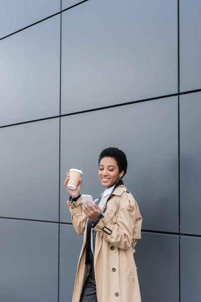 Pleased african american businesswoman in trendy trench coat messaging on mobile phone while standing near grey wall on street — Stock Photo