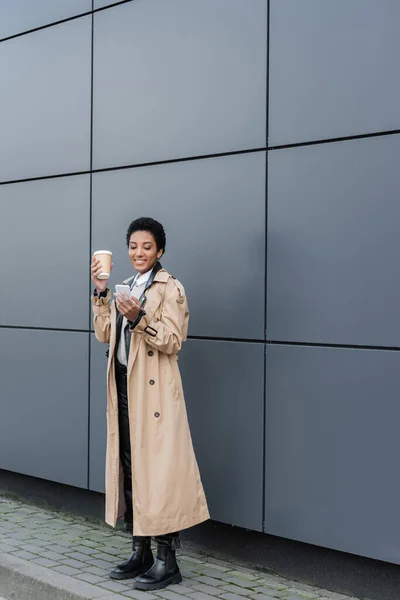 Piena lunghezza di felice e alla moda afroamericana donna d'affari in possesso di tazza di carta e messaggistica sul telefono cellulare vicino alla parete grigia — Foto stock