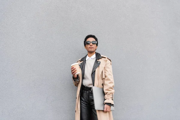 Модна афроамериканська бізнес-леді в бежевому пальто і сонцезахисних окулярах, що стоять з ноутбуком і кавою, щоб піти біля сірої стіни на міській вулиці — стокове фото