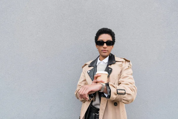 Donna d'affari afroamericana in occhiali da sole che tiene in mano la tazza di carta e controlla il tempo sull'orologio da polso mentre aspetta in strada — Foto stock