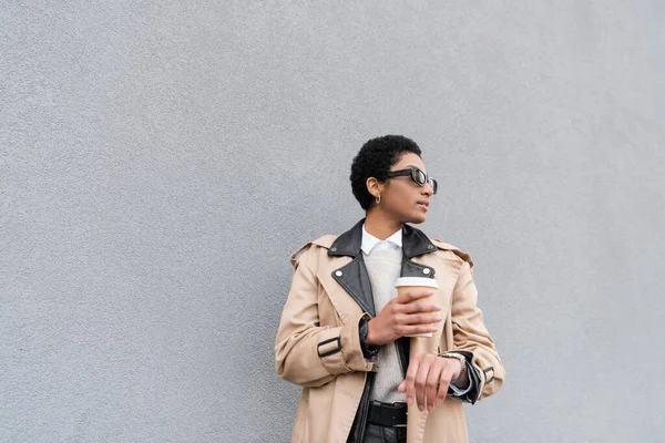 Young and fashionable african american businesswoman in sunglasses holding takeaway drink while waiting near grey wall on street — Stock Photo