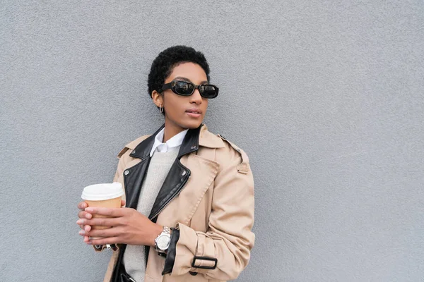 Femme d'affaires afro-américaine en trench coat beige et lunettes de soleil tenant tasse en papier et regardant loin près du mur gris en ville — Photo de stock