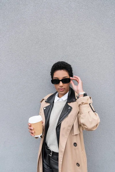 Donna d'affari afroamericana alla moda in trench che regola gli occhiali da sole e tiene la bevanda da asporto vicino al muro grigio sulla strada urbana — Foto stock