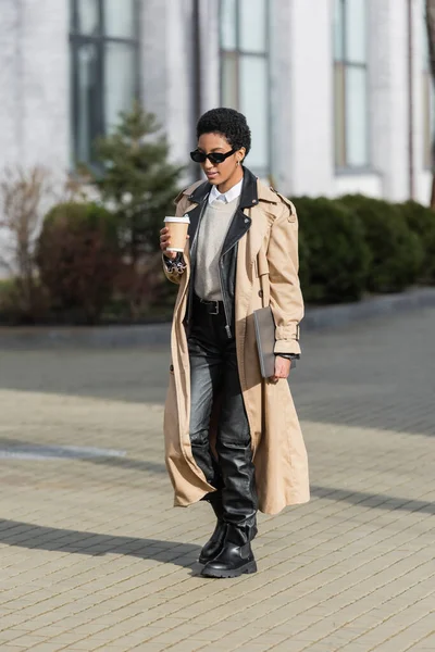 Comprimento total de mulher de negócios afro-americana com bebida takeaway e laptop andando em óculos de sol e casaco de trincheira na rua turva — Fotografia de Stock