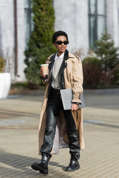 African american businesswoman in beige trench coat and sunglasses walking with disposable cup and laptop on blurred city street — Stock Photo