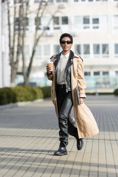 Full length of african american businesswoman in black leather pants and trench coat holding laptop and paper cup while walking on blurred street — Stock Photo