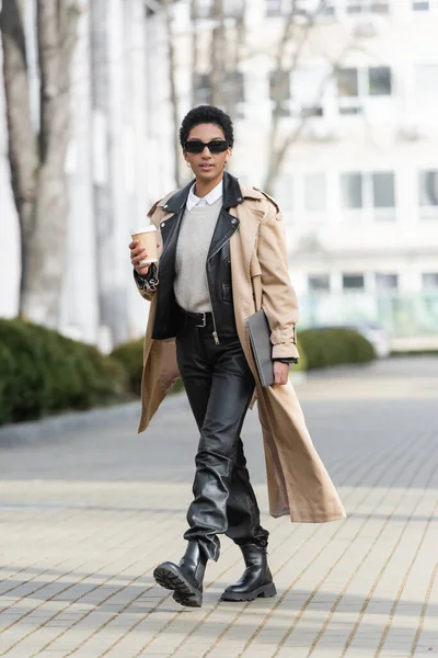Piena lunghezza della donna d'affari afro-americana alla moda in occhiali da sole che cammina con laptop e drink da asporto su strada sfocata in città — Foto stock