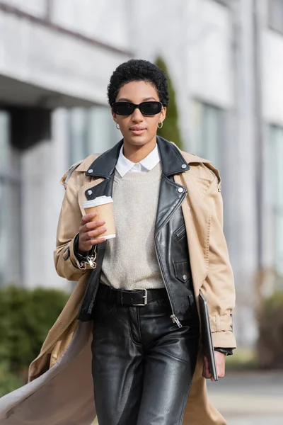 African american businesswoman in trench coat and black leather pants walking with coffee to go and laptop on blurred city street — Stock Photo