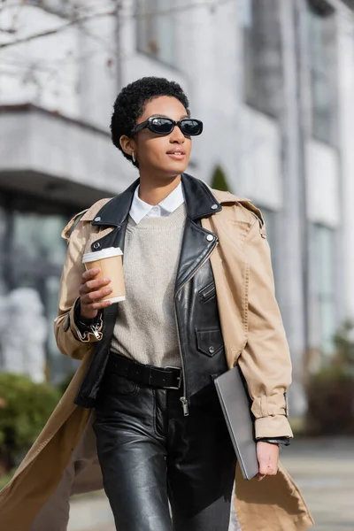 Stylish african american businesswoman in sunglasses holding laptop and disposable cup while walking and looking away on blurred street — Stock Photo