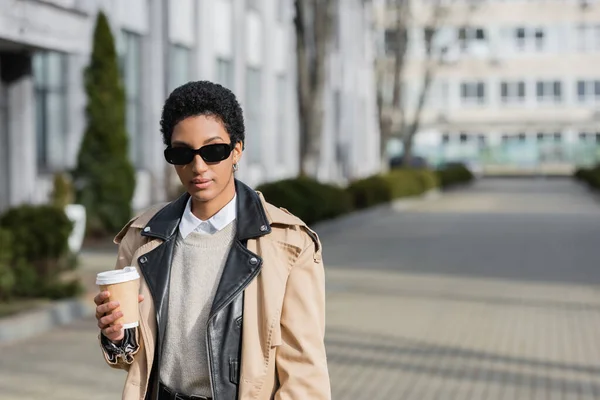 Молодая и стильная американская бизнесвумен в бежевом плаще и модных солнцезащитных очках, стоящих с бумажной чашкой на размытой улице — стоковое фото