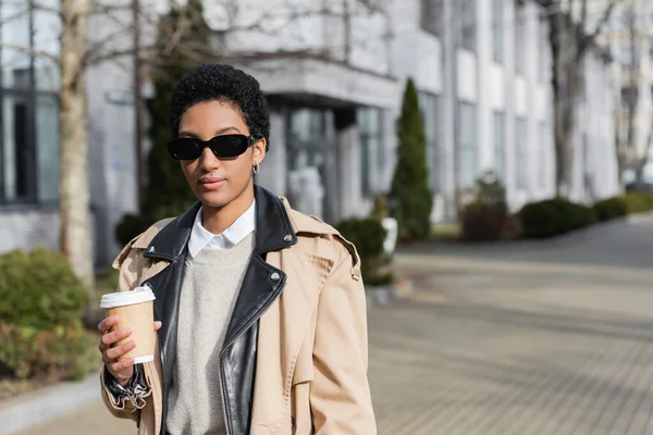 Jeune femme d'affaires afro-américaine avec des lunettes de soleil élégantes et un trench coat beige tenant une tasse en papier sur la rue floue de la ville — Photo de stock