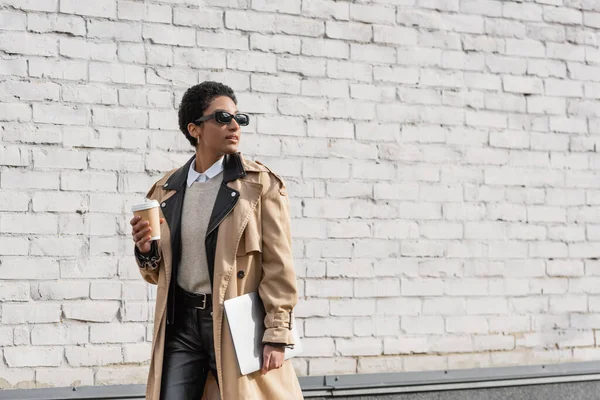 Femme d'affaires afro-américaine en lunettes de soleil et imperméable beige marche avec ordinateur portable et café pour aller le long du mur de briques sur la rue urbaine — Photo de stock