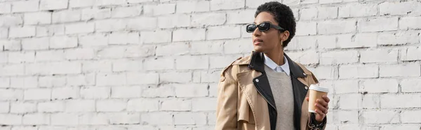 Donna d'affari afroamericana in occhiali da sole ed elegante trench che tiene il caffè per andare e guardare lontano vicino al muro di mattoni sulla strada, striscione — Foto stock